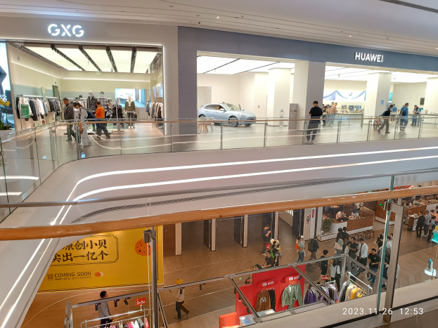 Ein Einkaufszentrumsinneres mit Menschen, die gehen, Glasgeländern, Bekleidungsauslagen, Informationsschildern, Säulen, Deckenleuchten und einem geparkten Auto im Hintergrund, mit einem GXG- und Huawei-Partnerschaftswasserzeichen.