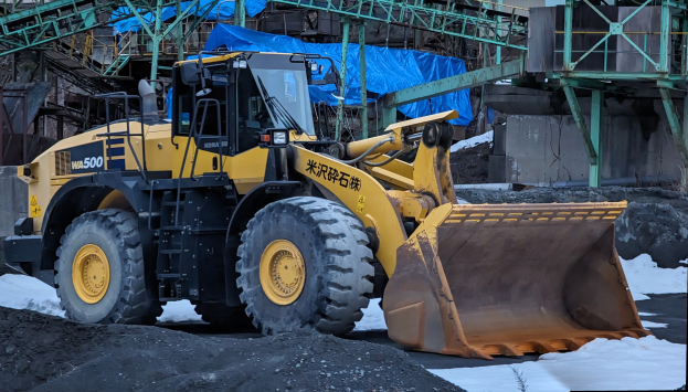 Ein großer gelber Radlader vor einem Gebäude geparkt, umgeben von Sand und Schnee, mit Eisenstangen und einer blauen Plane im Hintergrund.