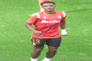 Emmanuel Adebayor, ein Mann in einem roten T-Shirt und schwarzen Shorts, läuft auf einem Fußballfeld mit zwei Personen im Hintergrund.