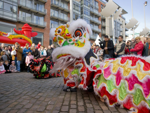 Ein lebendiges chinesisches Neujahrsfest in Amsterdam mit einem Löwen tanzen im Vordergrund, umgeben von Menschen und Gebäuden unter einem klaren blauen Himmel.
