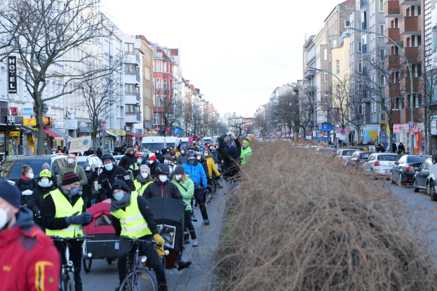 Eine große Gruppe von Menschen in Masken und Sicherheitswesten auf Fahrrädern eine von Bäumen gesäumte Straße mit Gebäuden, Laternenmasten, Texttafeln, Fahrzeugen, trockenem Gras und einem klaren blauen Himmel.