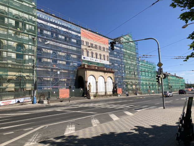 Großes modernes Gebäude im Bau in Berlin, umgeben von städtischen Elementen wie Straßenlaternen, Ampeln, Fahrzeugen, Fußgängern, Werbetafeln, Bäumen und einem bewölkten Himmel.