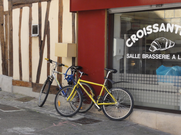 Zwei Fahrräder nebeneinander auf einem Gehweg vor einem Geschäft mit Glaswand und Text, mit einem Objekt an der Wand links von den Fahrrädern.