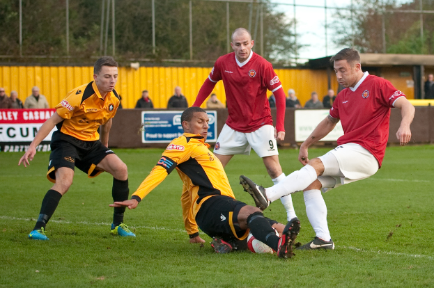 Spieler in blauen und roten Uniformen spielen ein Spiel auf einem grünen Feld mit einem Ball, während Zuschauer außerhalb des Feldes stehen und sie anfeuern, mit einem Baum und Himmel im Hintergrund.