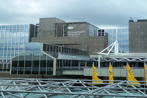 Frankfurt Airport in Frankfurt, Germany, a large building with glass walls and text, surrounded by yellow flags and iron rods, with a sky filled with white, fluffy clouds in the background.