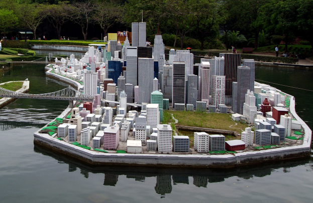 Ein Miniaturmodell einer Stadtlandschaft auf Wasser mit Gebäuden, Grünflächen, einer Brücke, Menschen und Bäumen unter einem Himmel-Hintergrund.