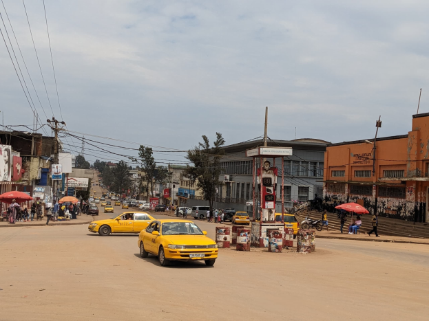 Ein gelbes Taxi fährt eine belebte Stadtstraße entlang, die von Gebäuden gesäumt ist, wo Fußgänger mit Regenschirmen auf dem Gehweg gehen, unter einem bewölkten Himmel, mit Strommasten und Drähten im Hintergrund.