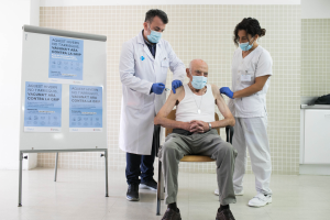 Ein älterer Mann sitzt in einem Stuhl in einer Krankenhausumgebung, mit zwei maskierten und handschuhgetragenen Ärzten, die neben ihm stehen, scheint eine Impfung zu erhalten, mit einer Tafel auf der linken und Gegenständen auf einer Plattform auf der rechten Seite.