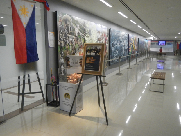 Langer Flur mit einer philippinischen Flagge an der Wand, Displaytafeln mit Text und Bildern, Ribbon-Ständer, Bänke, Deckenleuchten und ein Bildschirm im Hintergrund mit Museumsinformationen.