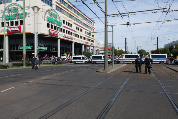 Eine Gruppe von Polizisten steht an der Straßenseite neben einer Tram, mit Fahrzeugen, Strommasten, Gebäuden mit Namensschildern, Bäumen und einem bewölktem Himmel im Hintergrund.
