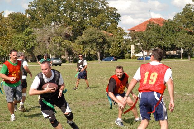 Menschen beim American-Football-Spielen auf einem grasbewachsenen Feld mit Bäumen, einem Haus und Autos im Hintergrund.