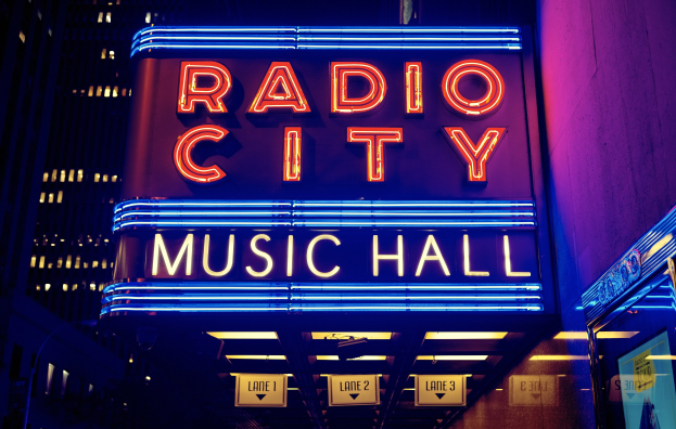 Das erleuchtete Radio City Music Hall in New York City, umgeben von hohen Gebäuden, mit einem "Radio City"-Schild und zusätzlichen Schildern an seiner Basis.