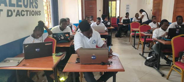 Eine Gruppe von Menschen sitzt an Schreibtischen mit Laptops in einem Klassenzimmer, mit Taschen auf dem Boden und Text an der linken Wand, was auf einen Online-Kurs in Ghana hinweist.