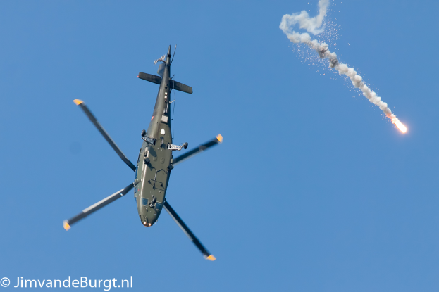 Ein Militärhubschrauber im Flug, der eine Rakete hält, die Rauch und Feuer ausstößt, mit Text in der linken unteren Ecke.