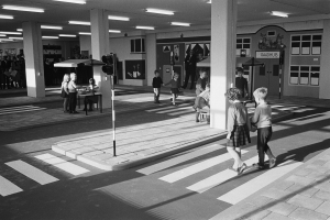 Schwarz-weiß-Foto von Kindern, die eine Straße an einer Ampel überqueren, mit Menschen auf Bänken, einem Verkehrssignalmast im Vordergrund und Säulen mit Deckenleuchten im Hintergrund.
