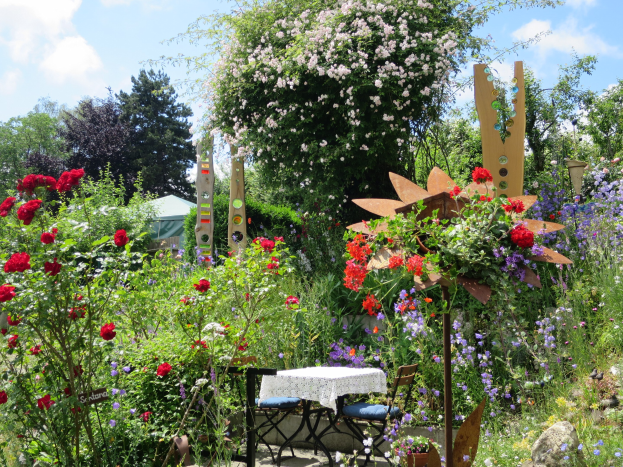 Ein Garten mit bunten Blumen und Pflanzen, der einen Tisch mit einer Decke und Stühlen zeigt, vor Bäumen und einem bewölkten Himmel beim Chelsea Flower Show.