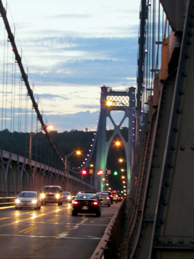 Fahrzeuge auf einer Brücke mit Lichtern, Pfählen, Hügeln und Himmel im Hintergrund.
