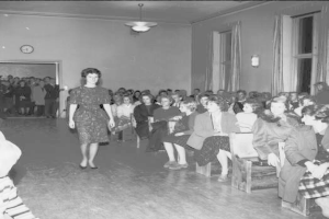 Eine Frau in einem schwarzen und weißen Kleid läuft über einen Laufsteg während einer Modenschau für Frauenwahlrecht, mit einer Menge, die auf Stühlen sitzt und einer Uhr und Lichtern an der umgebenden Wand.