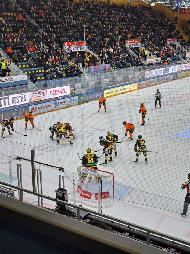Eine Gruppe von Menschen, die Hockey auf einer Indoor-Eisfläche mit einem Tor spielen, umgeben von einem Zaun, unter Stadionbeleuchtung, mit Zuschauern im Hintergrund und sichtbaren Bannern.