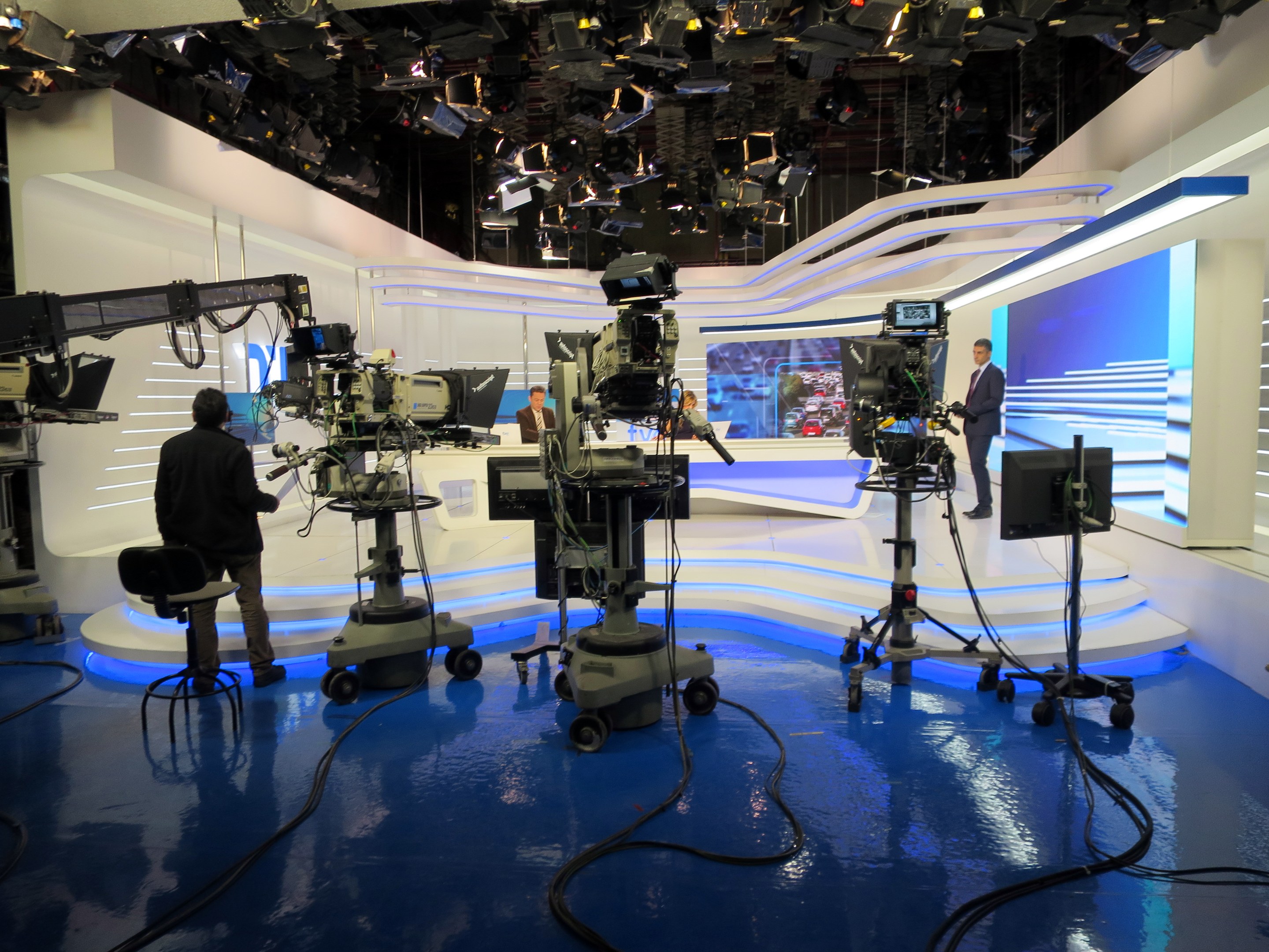 Ein TV-Studio mit mehreren Kameras auf dem Boden, einer sitzenden Person im Vordergrund und einigen stehenden Personen im Hintergrund, mit deckenmontierten Lichtern für eine Livesendung.