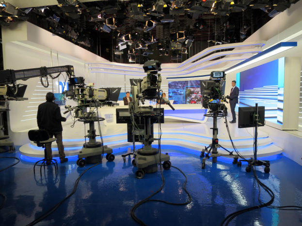 Ein TV-Studio mit mehreren Kameras auf dem Boden, einer sitzenden Person im Vordergrund und einigen stehenden Personen im Hintergrund, mit deckenmontierten Lichtern für eine Livesendung.