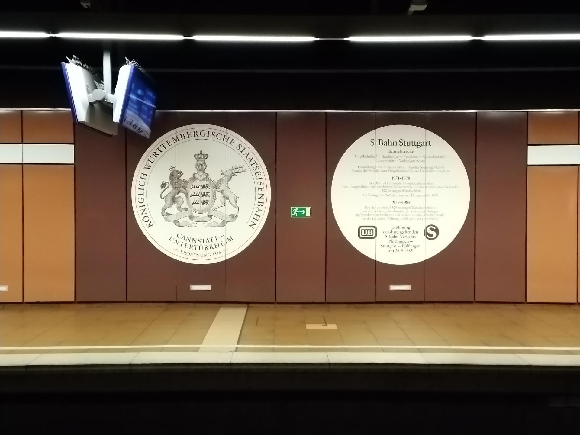 Ein Bahnsteig mit einem Schild, auf dem "S-Bahn Stuttgart" steht und einem Logo an der Wand, Deckenleuchten und ein paar Objekte auf der linken Seite des Bahnsteigs.