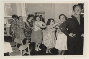 Eine Gruppe von Menschen, die in einem Raum mit Stühlen, einem tisch mit weißer Decke, einem Foto auf der Wand und Fenstern tanzen, gekleidet in 1950er-Jahre-Kleidung.