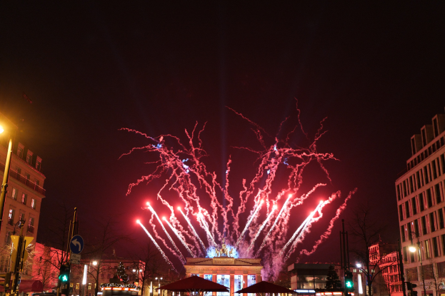 Eine belebte Stadtstraße während der Silvesterfeierlichkeiten in Berlin, voller Menschen, Fahrzeuge und festlicher Beleuchtung von Gebäuden und Feuerwerk.