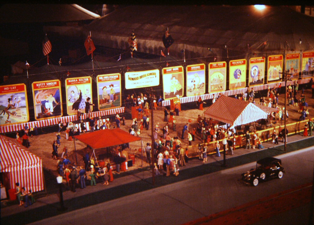 Ein altes Faschingsbild mit Menschen, einem Auto, Masten mit Fahnen, einem Zaun, einem Zelt, Brettern mit Bildern und Texten und einer Laterne.
