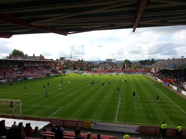 Ein Fussballspiel wird im St. Mary's Stadium gespielt, mit Zuschauern auf den Bänken und auf dem Rasen sitzend, einem Torpfosten links und Gebäuden, Bäumen und einem bewölkten Himmel im Hintergrund.