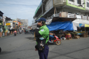 Ein Polizeibeamter in grüner und blauer Uniform und blauer Mütze steht auf der Straße und spricht in ein Handy, während Autos vorbeifahren, Stände mit Gegenständen rechts von ihm und ein bewölkter Himmel im Hintergrund.