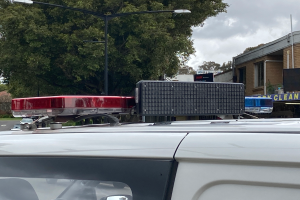 Ein Polizeiwagen mit eingeschaltetem Blaulicht steht am Straßenrand, umgeben von Gebäuden, Bäumen und Masten, mit Texttafeln in der Nähe, unter einem bewölkten Himmel, wahrscheinlich nach einer tödlichen Schulschießerei in Brisbane.