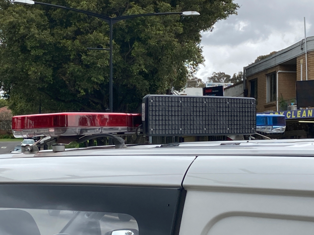 Ein Polizeiwagen mit eingeschaltetem Blaulicht steht am Straßenrand, umgeben von Gebäuden, Bäumen und Masten, mit Texttafeln in der Nähe, unter einem bewölkten Himmel, wahrscheinlich nach einer tödlichen Schulschießerei in Brisbane.