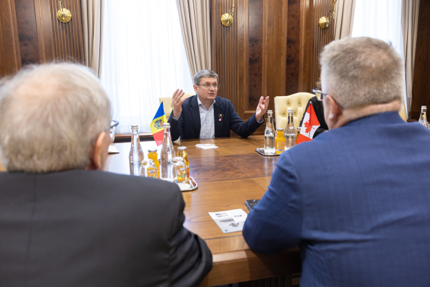 Drei Männer in einem formellen Setting sitzen um einen Holztisch mit Flaschen, Gläsern, Flaggen und Papieren, die in ein Gespräch vertieft sind.