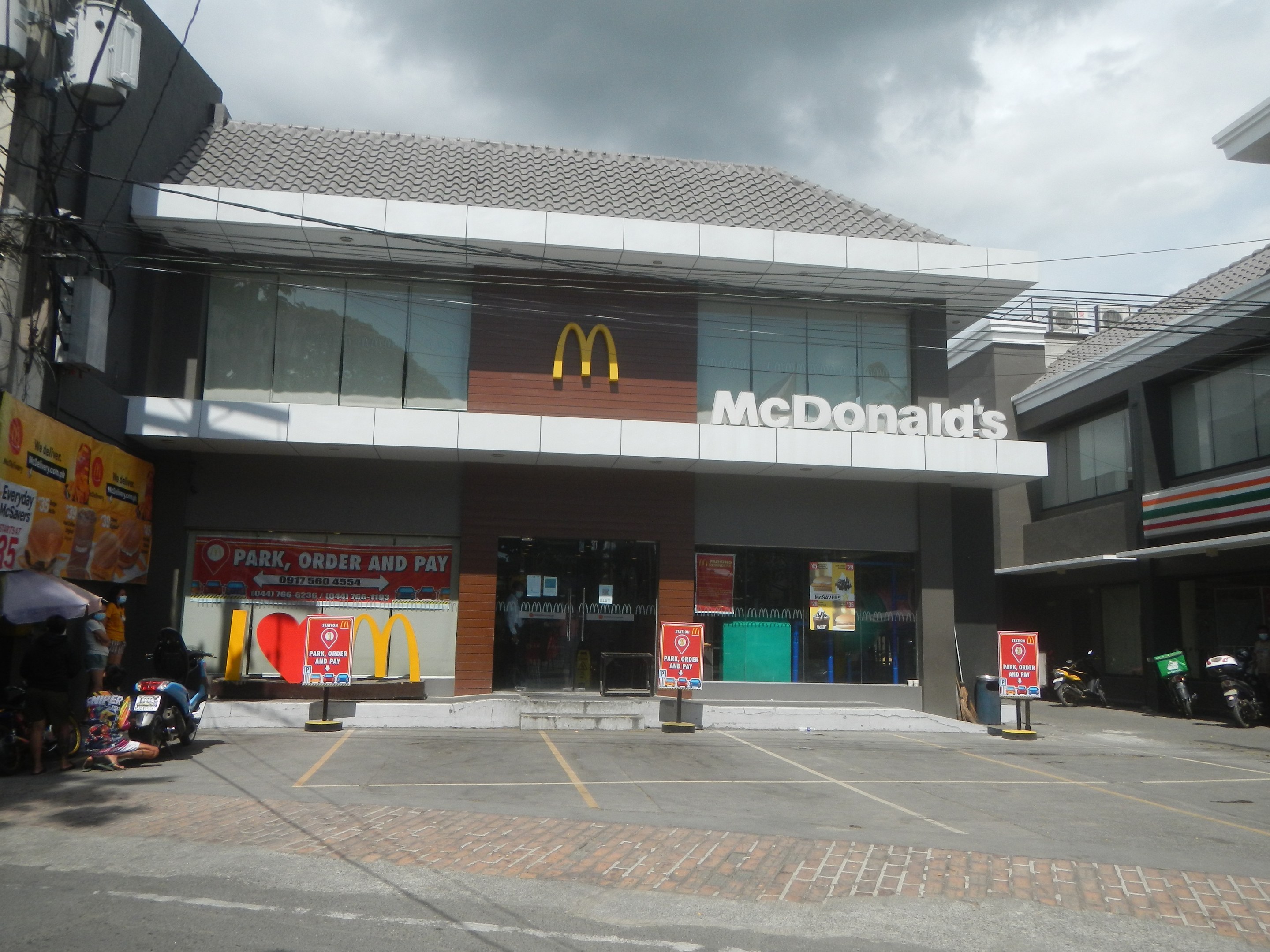 McDonald's Restaurant an einer Straßenecke mit Gebäuden, Motorrädern, Menschen, Schirmen, Werbetafeln, Strommasten, Kabeln und einem bewölkten Himmel im Hintergrund.