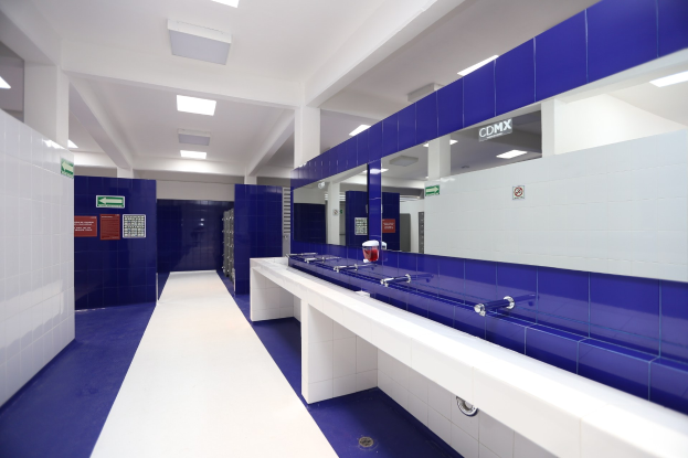 Öffentliche Toilette mit blauen Kachelwänden, weißen Arbeitsplatten, Waschbecken mit Wasserhähnen, einer Plattform mit einer Flasche und anderen Gegenständen, Informationsschildern an den Wänden, Deckenleuchten und tragenden Säulen.