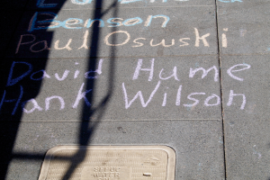 Ein Gehweg mit Kreide-Schrift, die wie ein Denkmal aussieht, ein Gullideckel unten und ein Schatten eines Baums auf der linken Seite.