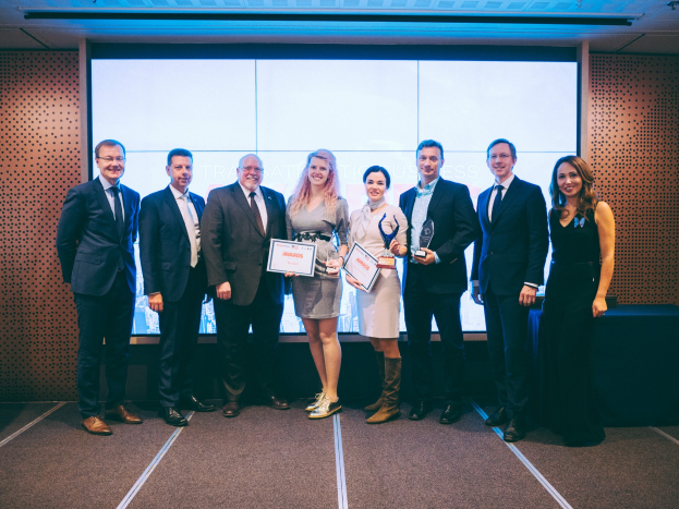 Eine Gruppe strahlender Menschen mit Urkunden, die vor einem Bildschirm stehen, mit einem Tisch und einem wandmontierten Objekt im Hintergrund, Gewinner der International Business Awards 2018.