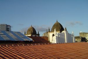 Eine Stadtansicht mit einigen Gebäuden im Vordergrund und einem blauen Himmel im Hintergrund, auf dessen Dach sich Solarpanels befinden.
