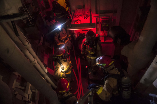 Feuerwehrleute in Helmen und Sauerstoffflaschen stehen in einem schwach beleuchteten Raum mit Rohren an den Seiten, bereit für einen Rettungseinsatz in der Nähe von Maschinen und anderen Gegenständen.