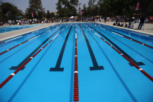 Großes Schwimmbad mit Bahnentrennung, umgeben von Menschen mit Taschen und Gegenständen, Zelten, Bannern, Masten, einem Schild und einem bewölktem Himmel im Hintergrund.