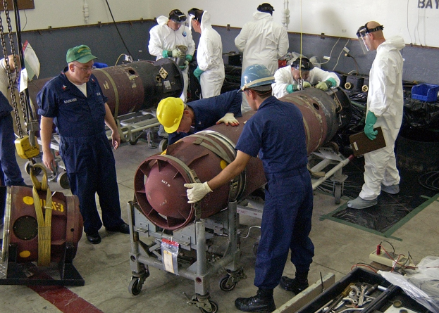 Männer in Helmen und Handschuhen, die eine große Bombe in einer Fabrik mit Maschinen und Wandtext entschärfen.