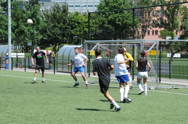 Menschen in Kleidung beim Fußballspielen auf einem Feld mit einem Zaun und einem Tor in der Mitte, einem Pfahl und einem Fußball oben links und Bäumen und Gebäuden im Hintergrund.