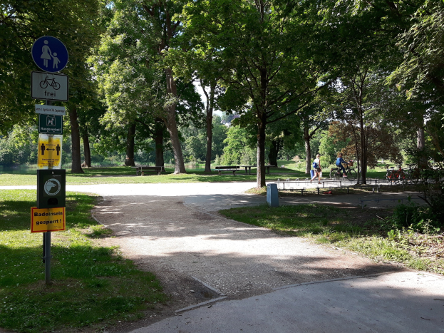 Ein asphaltierter Parkweg, gesäumt von Bäumen und Gras, mit Schildern an einem Pfahl auf der linken Seite und Radfahrern im Hintergrund.