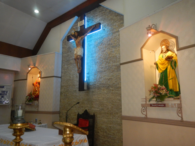 Das Innere einer Kirche mit einem Kreuz an der Wand, einer Jesus-Statue am Kreuz, einem Tisch mit einer Decke und verschiedenen Gegenständen, einem Stuhl, einem Mikrofon mit Ständer, einer Tafel mit Text auf der linken Seite, Deckenlampen und Blumensträußen auf der rechten Seite.
