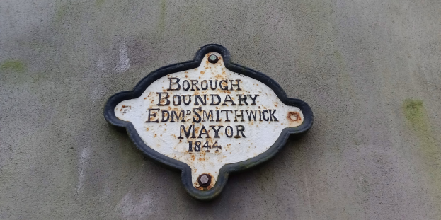 Metallschild an der Seite eines Gebäudes mit schwarzem Hintergrund und weißer, fetter, zentrierter Schrift, die "Borough Boundary Edm Smithwick Mayor" liest.