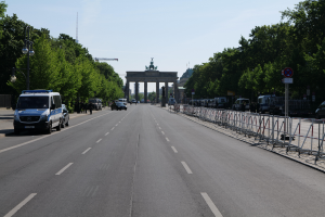 Ein Polizeiwagen steht auf der Seite einer vielbefahrenen Straße vor dem Brandenburg-Tor in Berlin, Deutschland, mit Barrieren, Schildern, Bäumen und Laternenmasten im Hintergrund und einer bewölkten Himmel.