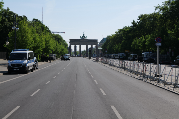 Ein Polizeiwagen steht auf der Seite einer vielbefahrenen Straße vor dem Brandenburg-Tor in Berlin, Deutschland, mit Barrieren, Schildern, Bäumen und Laternenmasten im Hintergrund und einer bewölkten Himmel.