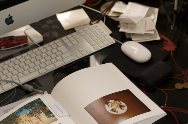 Ein Schreibtisch mit einem Monitor, Tastatur, Buch, Papieren, Maus, Münzen und einem Spielzeugauto.