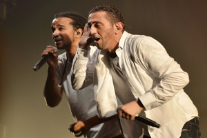 Zwei Männer in legerer Kleidung performen auf der Bühne Seite an Seite, jeder hält ein Mikrofon.